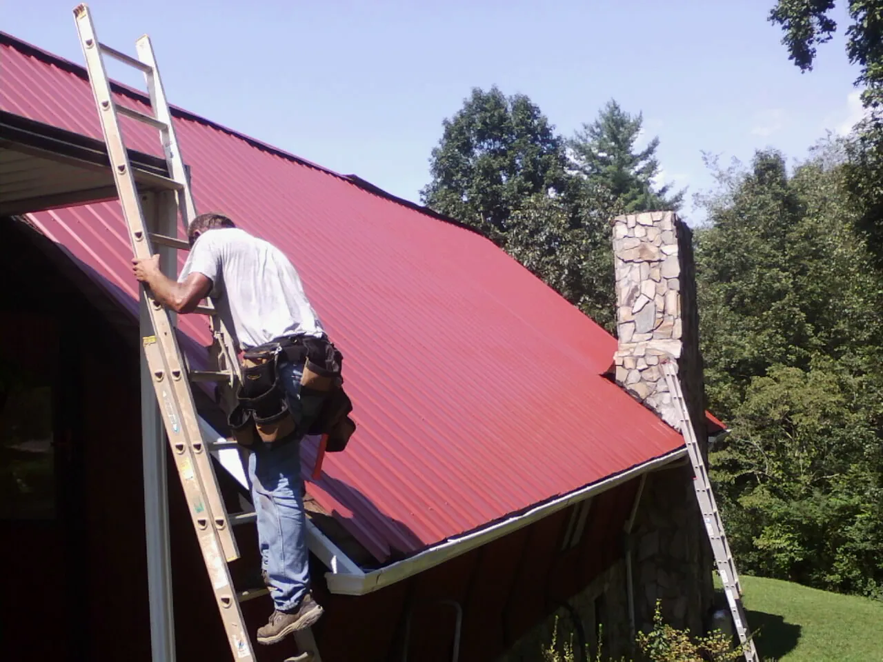 Roofer on a ladder beside a red metal roof and stone chimney — Soffit & Fascia Repair by Roofing Brothers in Piscataway