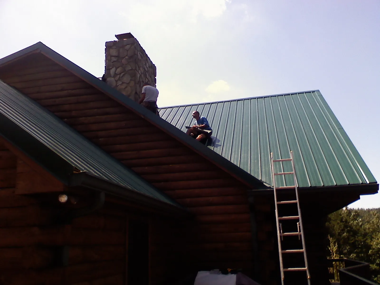 Roofers installing green metal panels beside a stone chimney on a log home in Piscataway